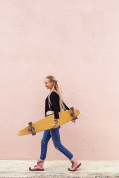 City portrait of positive young female  holding skateboard. Pink wall on background