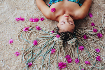 Young woman wearing braids hair style lying on sea shore with pink flowers in her hair