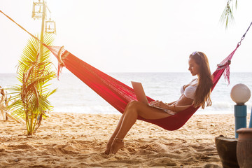 Young woman working on laptop seating in hammock under palm trees on tropical beach