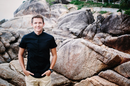 Outdoor Portrait of Young smiling businessman wearing black shirt standing on ricks sea shore
