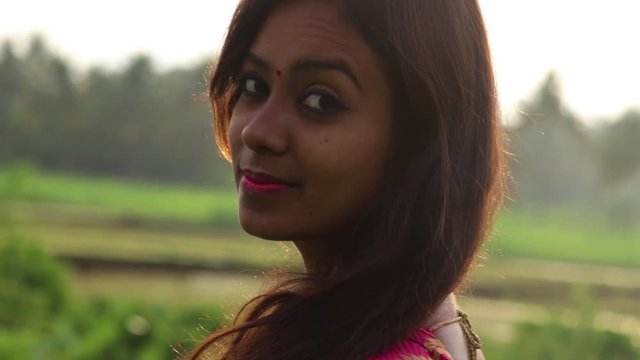 Closeup of a Indian woman in traditional sari dress standing in a grass field, turns around to look at the camera and smile.