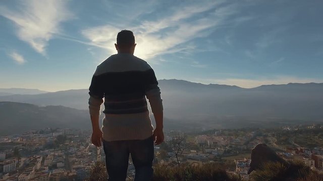 Camera follows young Moroccan man walking up  on top of mountain summit, Chefchaouen Morocco, sunny day