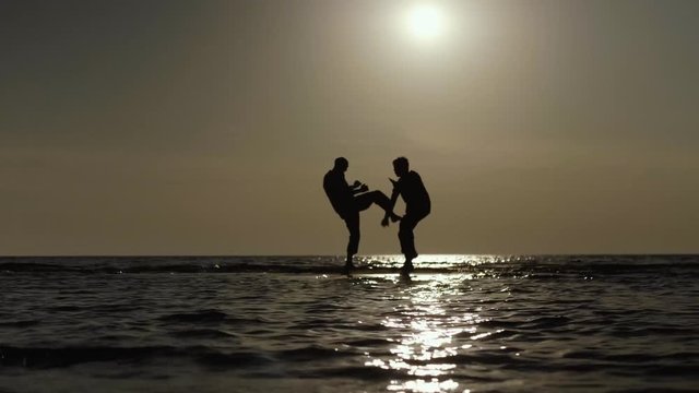 Two martial artists practicing light sparring on the beach at sunset.