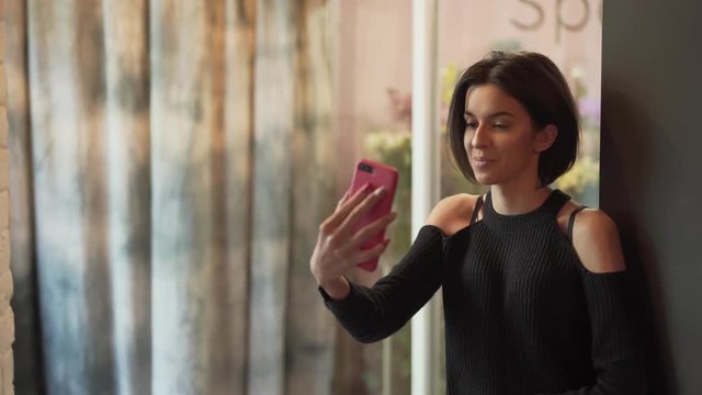 Beautiful girl is standing near the showcase in a flower shop and talking on the phone in a video chat. Girl has very expressive face features and movements.