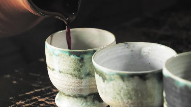Slow Motion of red wine being poured from an ancient wine jug into a row of colorful wine goblets.