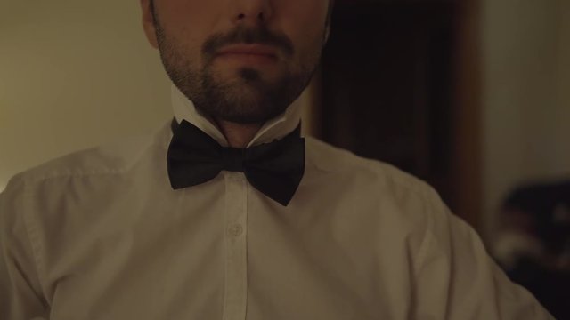 A Man In White Shirt And Black Batterfly Sits On A Bed With Close Eyes.Man sitting on couch at home.Tie Butterfly close-up.Black bow tie close up.Young handsome male sitting at the end of a bed.