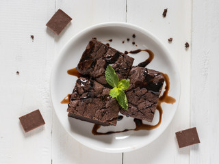 A plate of brownies with chocolate sauce on white wooden background, mint leaf on top, top view