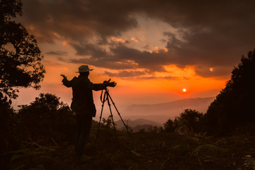 The male silhouette photographer stands on the mountain behind the sunset.