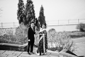 Elegant and fashionable indian friends couple of woman in saree and man in suit posed on stairs against lake.