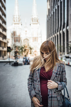Pregnant girl blonde in a burgundy dress in shape and a gray jacket. In the big city. Harm to the environment. Long hair. 9 months in anticipation. High-rise buildings. Urbanism. Happiness to be mom.
