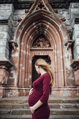 Pregnant girl blonde in burgundy dress. In the big city. Long hair. 9 months waiting. Happiness to be a mother. Against the backdrop of a beautiful old building.