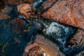 Smooth stones in spring water close-up. Clean water flow among red and orange stones. Colorful natural background of mountain spring stream with copy space. Beautiful texture of creek with wet stones.