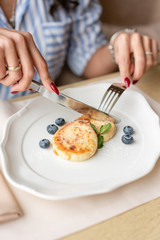 Beautiful young woman cuts pancakes with a knife. Light breakfast with coffee near window in a cafe.