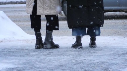 Young fashionable people girl friends standing on street intersection during snowfall. Closeup on boots. Slow motion.