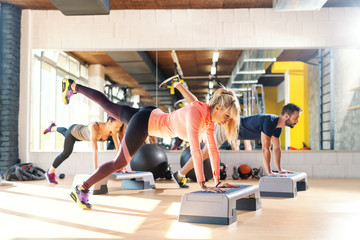 Group of people with healthy habits doing exercises for legs on steppers. Gym interior. In background mirror with their reflection.