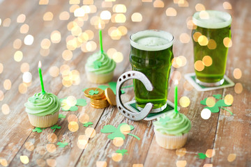 st patricks day, holidays and celebration concept - glass of green beer, cupcakes with candles, horseshoe and gold coins on wooden table