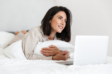 Photo of pretty woman 30s using laptop, while lying in bed with white linen in bright room