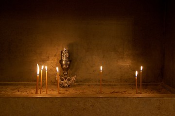 Lit candles candle light and a golden vigil oil lamp kept permanently burning placed on sand in a church of a Greek monastery in darkness in Greece - obrazy, fototapety, plakaty