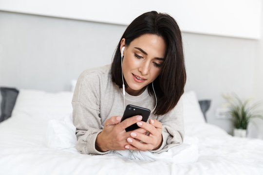 Image of beautiful woman 30s wearing earphones holding cell phone, while lying in bed in bright room