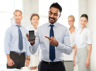 business, technology and people concept - smiling indian businessman pointing finger to blank screen of smartphone over colleagues on office background
