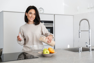 Image of brunette woman 30s making breakfast with oatmeal and fruits, while standing in modern kitchen at home - Powered by Adobe