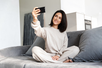 Photo of smiling woman 30s taking selfie on cell phone, while sitting on couch in bright apartment