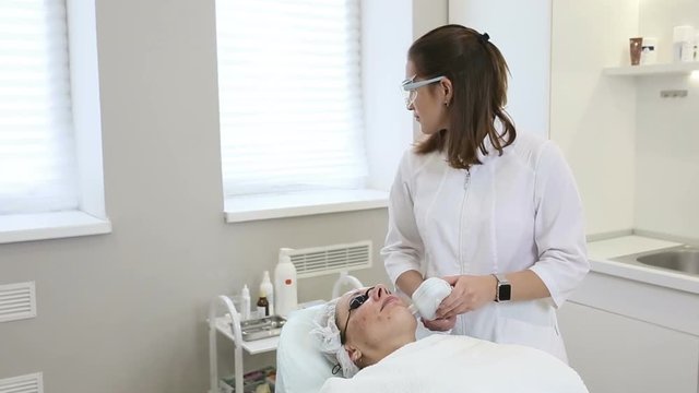 Woman in black glasses getting photo rejuvenation procedure in cosmetology clinic or beauty salon. Beautician doing facial procedure, Skin care concept for middle-aged woman.