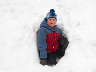 Fototapeta na wymiar Little boy is sitting at the entrance to a cave dugged by him in the snow. Portrait. Snowy winter concept.
