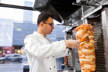 fast food and cooking concept - chef slicing doner meat from rotating spit at kebab shop