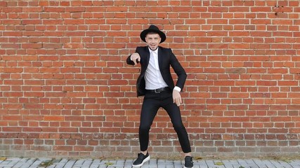 Young cheerful caucasian man in a hat and black suit dances against a brick wall and takes off his hat, copy space, slow-mo