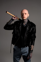 bald tattooed man in leather jacket holding baseball bat and looking at camera isolated on grey