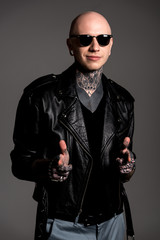 Obraz premium smiling tattooed man in leather jacket and sunglasses pointing at camera isolated on grey