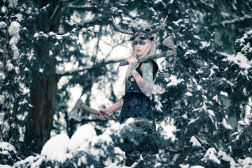 Beautiful warrior woman in image of viking with horned helmet and two axes in winter snowy forest.