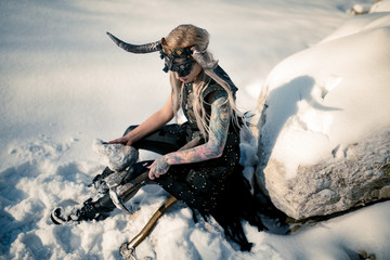 Warrior woman in image of viking with horned helmet and ax sits on snow next to big stone. - Powered by Adobe