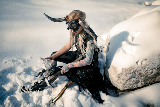 Warrior woman in image of viking with horned helmet and ax sits on snow next to big stone.