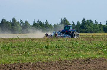 Fototapeta premium Blue tractor plowing the green field with an iron harrow in the plowing season with dust swirling