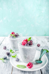 Summer healthy breakfast dessert in glass, overnight vegan chia pudding with non-dairy milk, frozen raspberry, blackberry and blueberry, on light blue and wooden background copy space