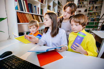 Four schoolchildren of different ages: first grader, preteen, teenager, adult female student or teacher learning together in library. Teaching children to do common project concept.