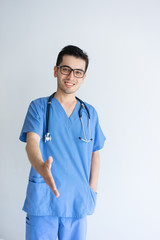 Smiling young male doctor extending arm for handshake. Handsome guy standing and wearing blue medical uniform. Medical offer concept. Isolated front view on white background.