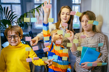Four different ages pupils with adult female student learning something from colorful stickers glued on glass wall during collaborative process.