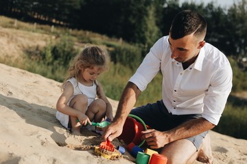 Happy caucasian family in white dress, dad playing with little girl with sand toys playing in sandbox at summer playground.