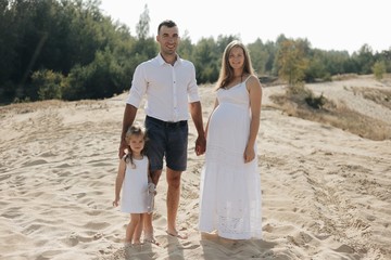Portrait of caucasian family with baby in white clothes are standing on sands dune. Pregnant mpther. Newborn