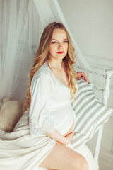 Naklejka premium Pregnant woman in dress holds hands on belly on a white background. Pregnancy, maternity, preparation and expectation concept. Close-up, copy space, indoors. Beautiful tender mood photo of pregnancy