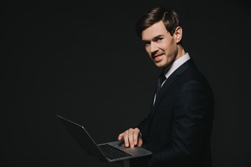 cheerful businessman in suit using laptop isolated on black