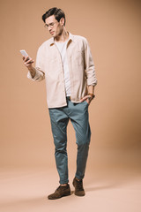 handsome man in glasses looking at smartphone while standing on beige background