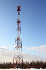 Big tower with a cellular antenna on a snowy field against the forest and blue sky on a winter day