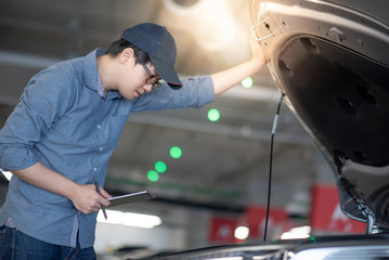 Asian auto mechanic holding digital tablet checking car engine under the hood in auto service garage. Mechanical maintenance engineer working in automotive industry. Automobile servicing and repair