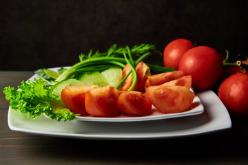 sliced tomatoes cucumbers lying on a white plate decorated with green onions standing on a dark wooden table