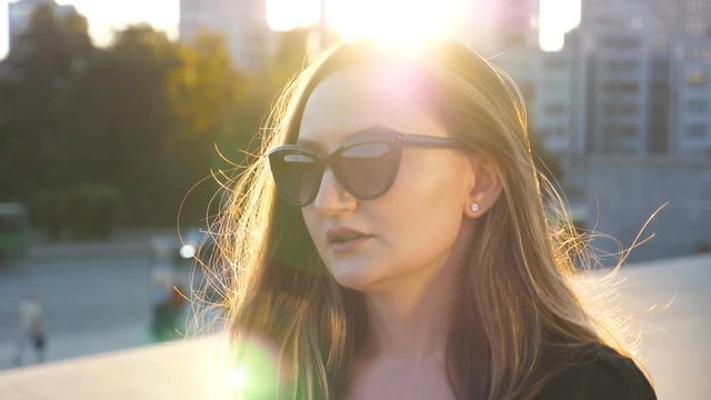 Portrait of young businesswoman in sunglasses walking in city street with sun flare at background. Face of attractive business woman commuting to work. Sunset light illuminates hair of girl. Close up