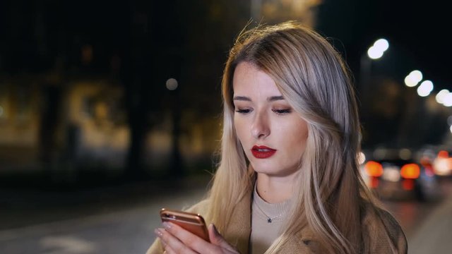 Attractive woman using smartphone during walk on streets of night town. Girl ordering taxi. Online service taxi order mobile application. Taxi online, cityscape.